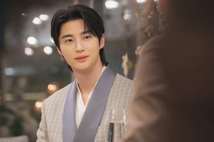 Seorang aktor mengenakan jas abu-abu duduk di meja makan dengan latar pencahayaan hangat dalam adegan drama Korea Perfect Crown.