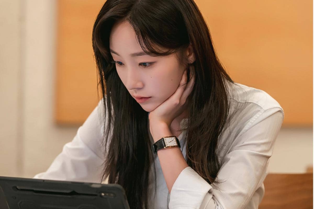Seorang wanita berambut panjang mengenakan kemeja putih sedang menatap layar tablet dengan ekspresi serius di ruangan dalam.