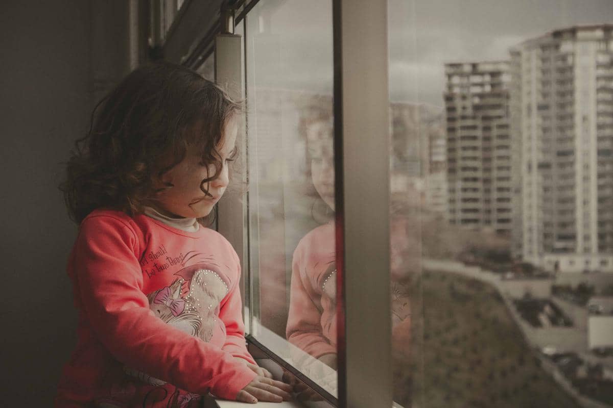 Seorang anak kecil berambut ikal mengenakan baju merah muda berdiri di dekat jendela sambil menatap keluar dengan ekspresi tenang.