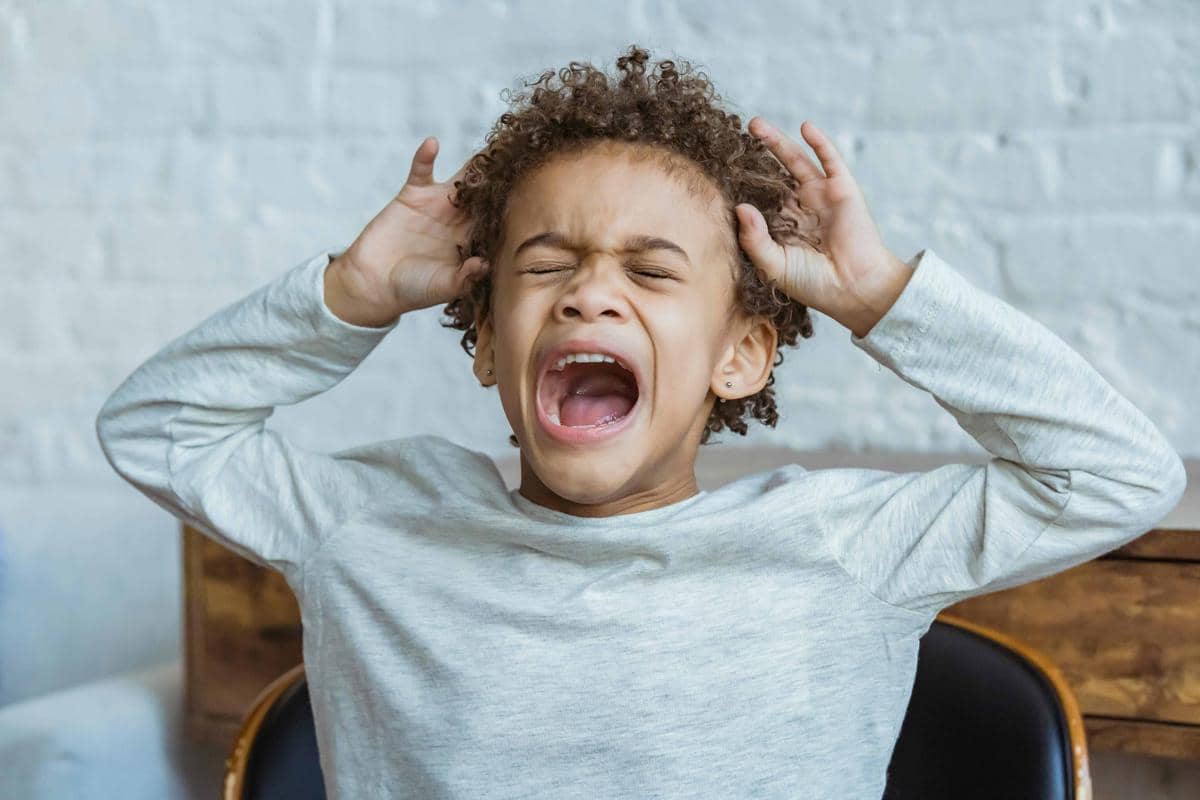 Anak kecil mengenakan kaus abu-abu berteriak sambil menutup telinga dengan kedua tangan di dalam ruangan.