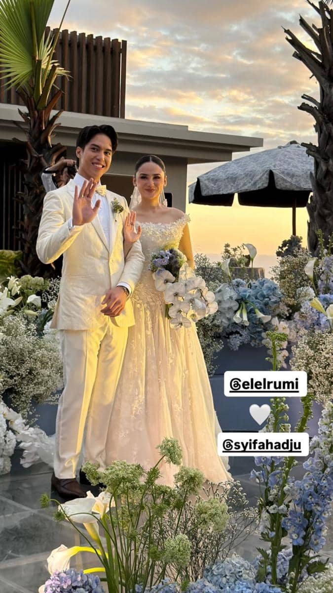 Pasangan pengantin berpose di resepsi pernikahan dengan latar bunga dan langit senja di Bali, mengenakan busana pengantin putih elegan.