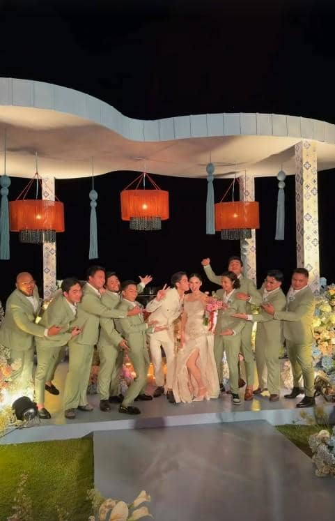 Pasangan berpakaian formal berpose di panggung pernikahan malam hari bersama rombongan pria bersetelan hijau pastel yang tampak ceria.