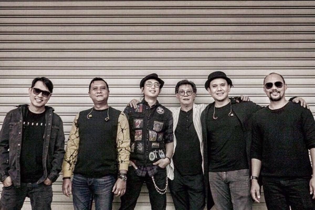 Enam pria berdiri berdampingan sambil tersenyum di depan pintu besi bergaris horizontal dengan gaya kasual dan santai.
