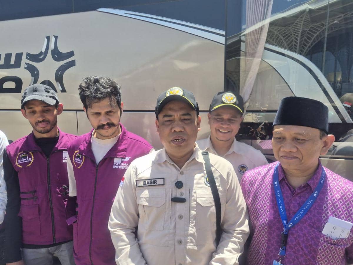 Beberapa petugas dan jemaah haji berdiri bersama di depan bus di Medinah, mengenakan seragam dan pakaian khas haji di bawah sinar matahari.