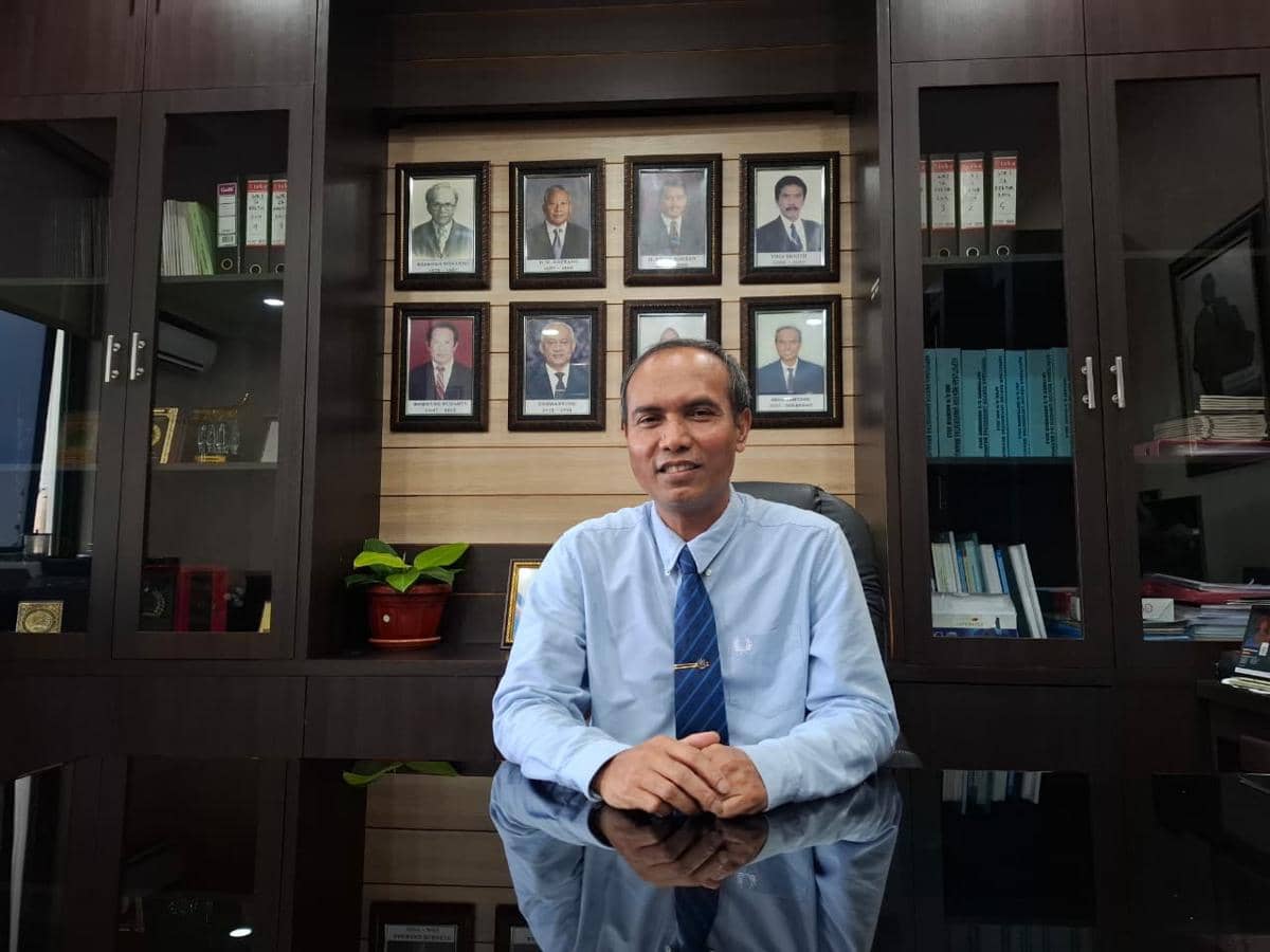 Seorang pria mengenakan kemeja biru dan dasi duduk di meja kantor dengan latar belakang rak buku dan foto-foto tokoh di dinding.