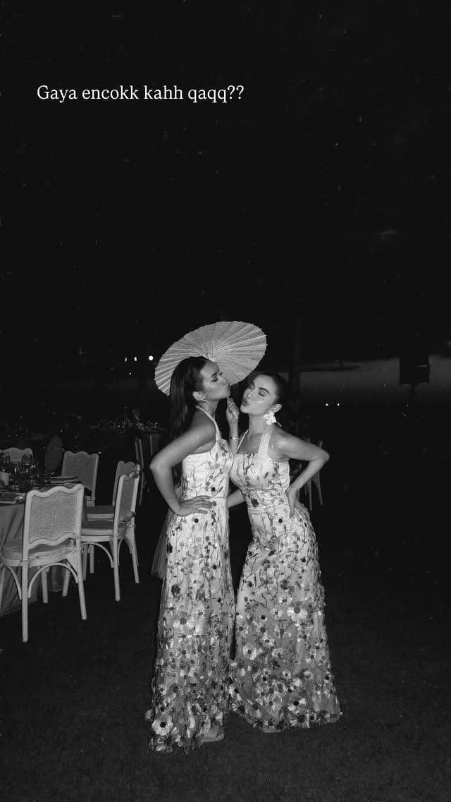 Dua wanita mengenakan gaun panjang bermotif bunga berpose bersama di acara malam hari dengan latar meja dan kursi pesta.
