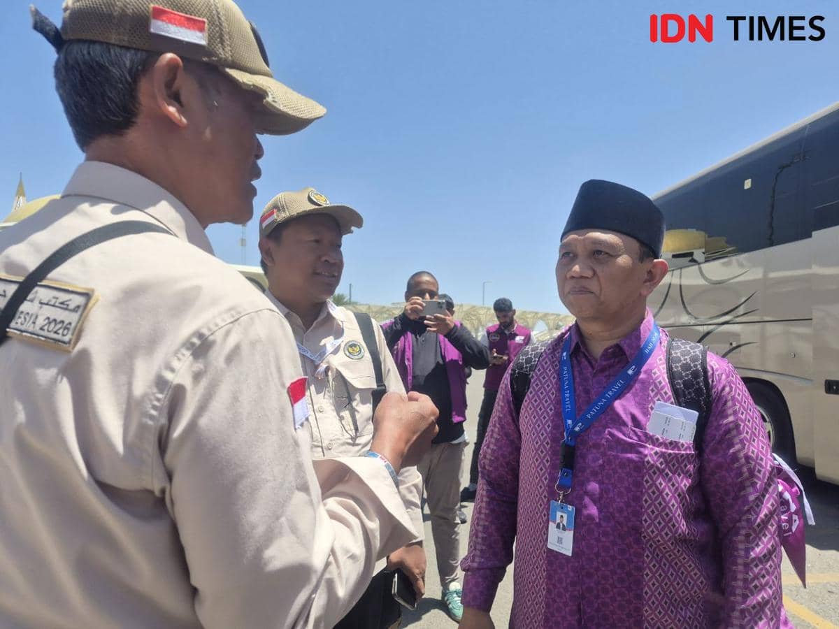 Petugas PPIH berbicara dengan jemaah haji khusus yang baru tiba, dengan latar belakang bus dan cuaca cerah di area keberangkatan.