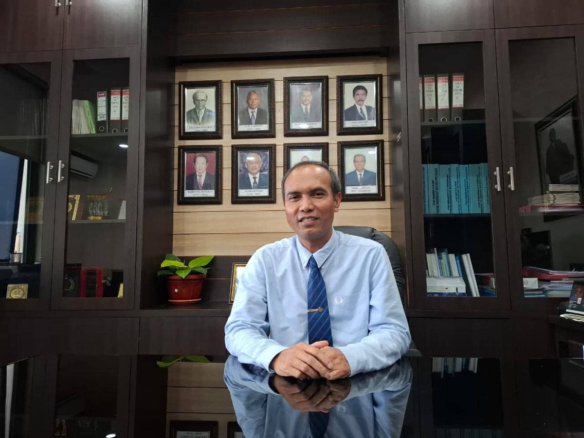 Seorang pria mengenakan kemeja biru dan dasi duduk di meja kantor dengan latar belakang lemari dan foto-foto tokoh di dinding.
