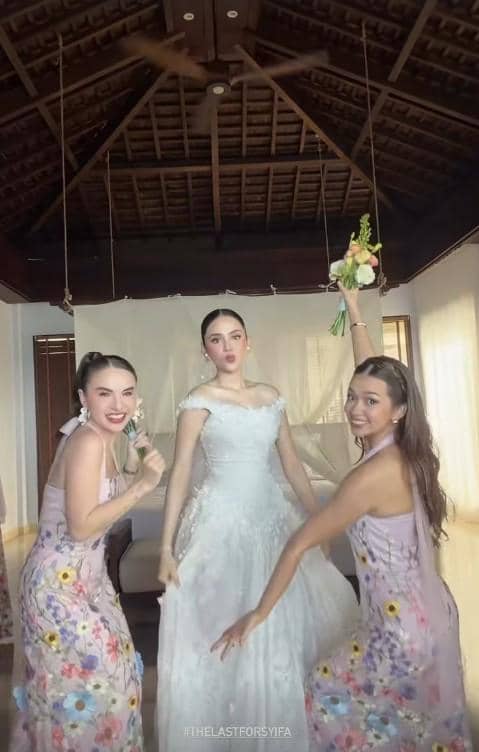 Tiga wanita berpose ceria di dalam ruangan, satu mengenakan gaun pengantin putih dan dua lainnya memakai gaun ungu bermotif bunga.