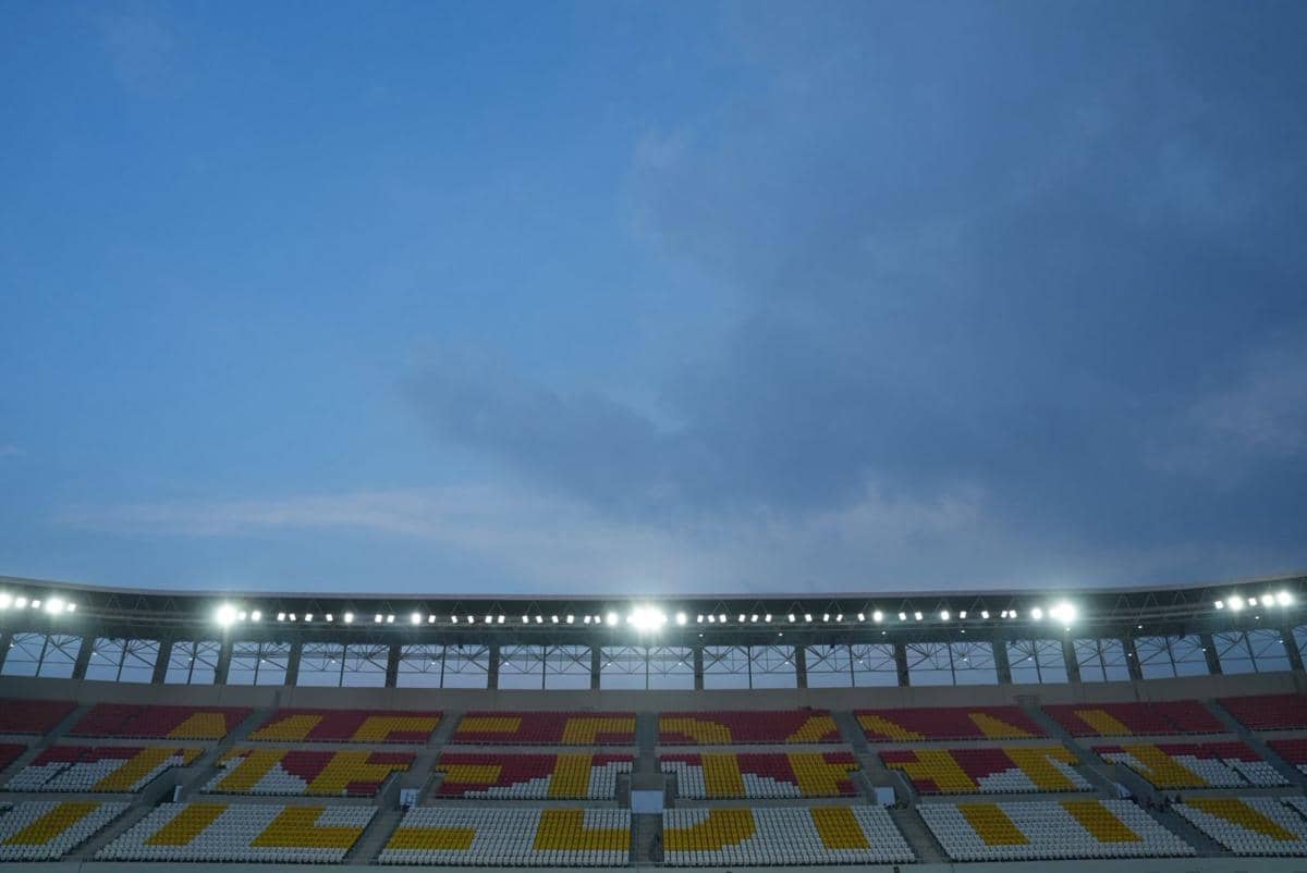 Kursi tribun Stadion Teladan Medan berwarna merah, kuning, dan putih dengan lampu stadion menyala di bawah langit senja.