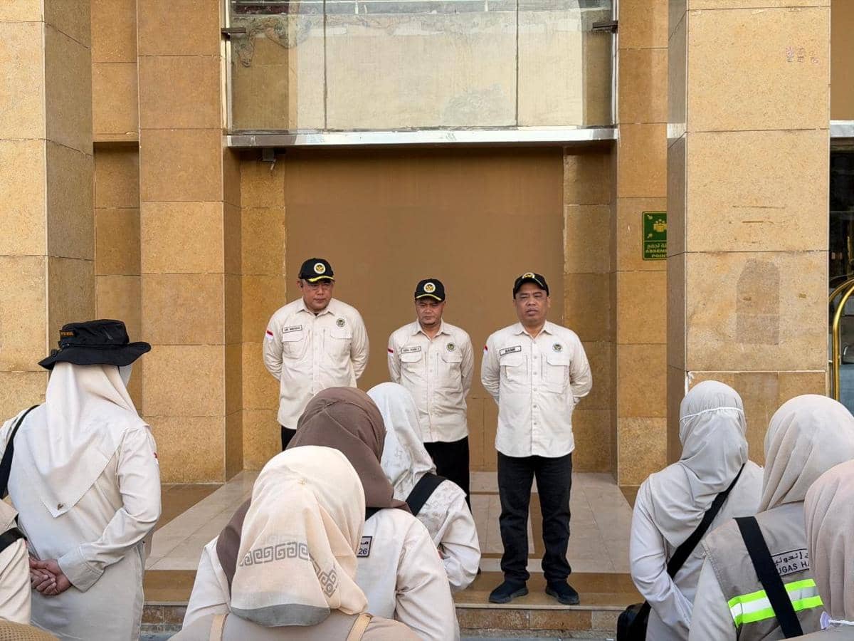 Kadaker Bandara Abdul Basir memimpin apel pagi petugas PPIH 2026 di depan gedung berwarna cokelat bersama dua petugas lainnya.