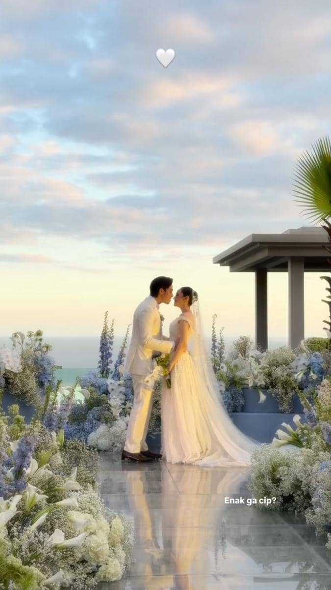Sepasang pengantin mengenakan busana putih berciuman di altar pernikahan outdoor dengan dekorasi bunga dan latar langit senja di Bali.