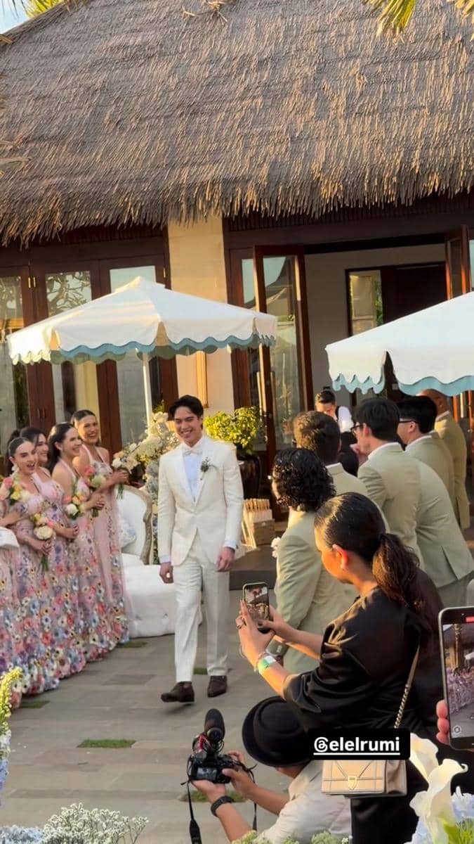 Seorang pria mengenakan setelan jas putih berjalan di area resepsi pernikahan outdoor di Bali dengan tamu dan pengiring pengantin di sekitarnya.