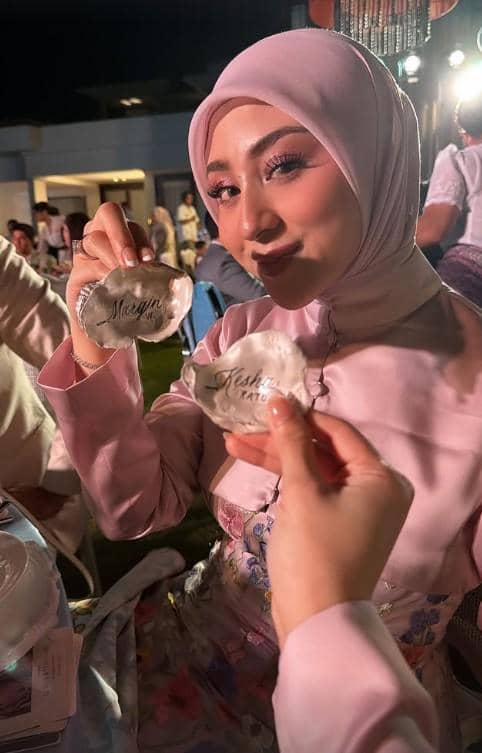Seorang wanita berhijab mengenakan busana berwarna pink memegang dua cangkang kerang bertuliskan nama di sebuah acara malam hari.