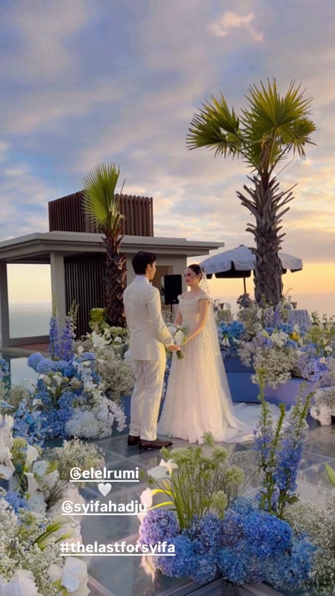 Pasangan pengantin berdiri di pelaminan outdoor dengan dekorasi bunga biru dan putih di Bali saat matahari terbenam.
