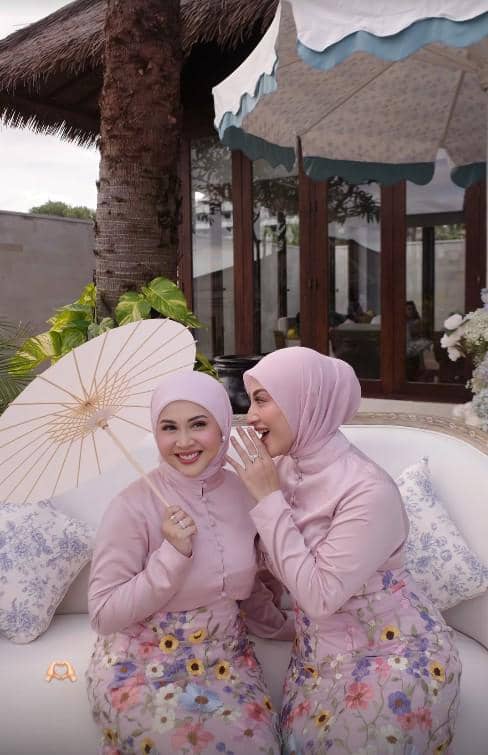 Dua perempuan berhijab mengenakan kebaya berwarna pink dan rok bermotif bunga duduk bersama sambil tersenyum di area outdoor bernuansa tropis.