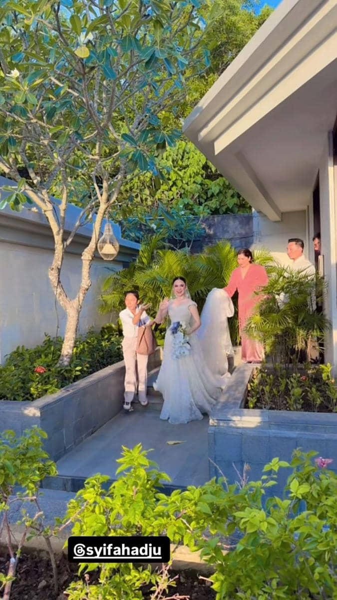 Seorang pengantin wanita mengenakan gaun putih berjalan di taman vila Bali ditemani keluarga, dikelilingi pepohonan hijau dan cahaya sore.