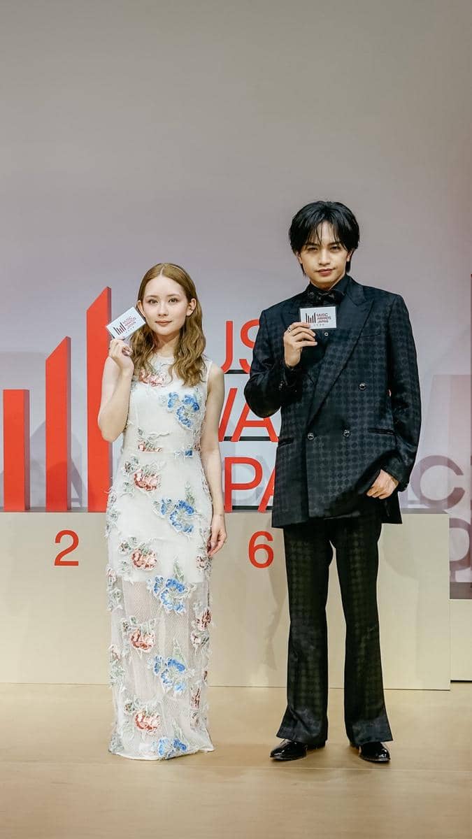 Dua duta Music Awards Japan 2026 berpose di atas panggung sambil memegang kartu acara, mengenakan busana formal elegan.