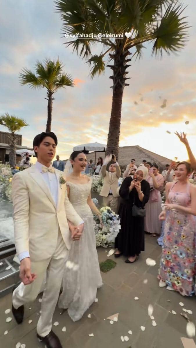 Pasangan pengantin berjalan bergandengan tangan di resepsi pernikahan outdoor dengan latar pohon palem dan tamu yang melempar bunga.