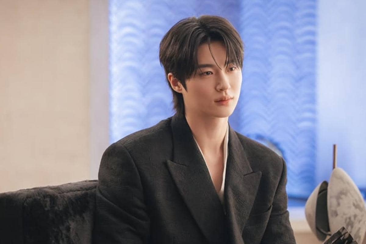 Seorang pria berambut hitam mengenakan jas hitam duduk di ruangan dengan latar tirai biru dalam cuplikan drama Korea Perfect Crown.
