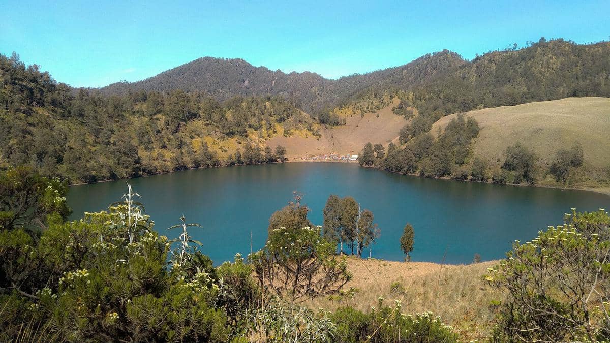 Danau Ranu Kumbolo dengan air biru jernih dikelilingi perbukitan hijau dan pepohonan di kawasan pegunungan Semeru.