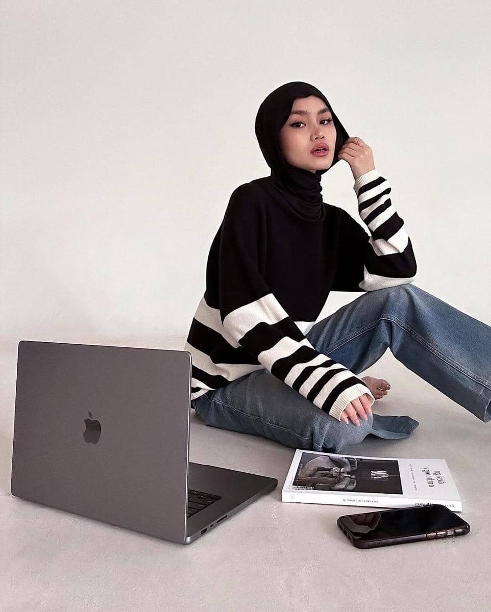 Seorang wanita berhijab mengenakan sweater hitam bergaris putih dan celana denim duduk di lantai dengan laptop, buku, dan ponsel di depannya.