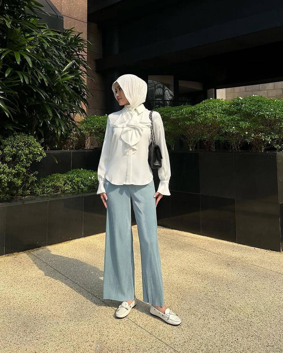 Seorang wanita berhijab mengenakan atasan putih dengan detail pita besar dan celana panjang biru muda, berpose di area luar gedung modern.