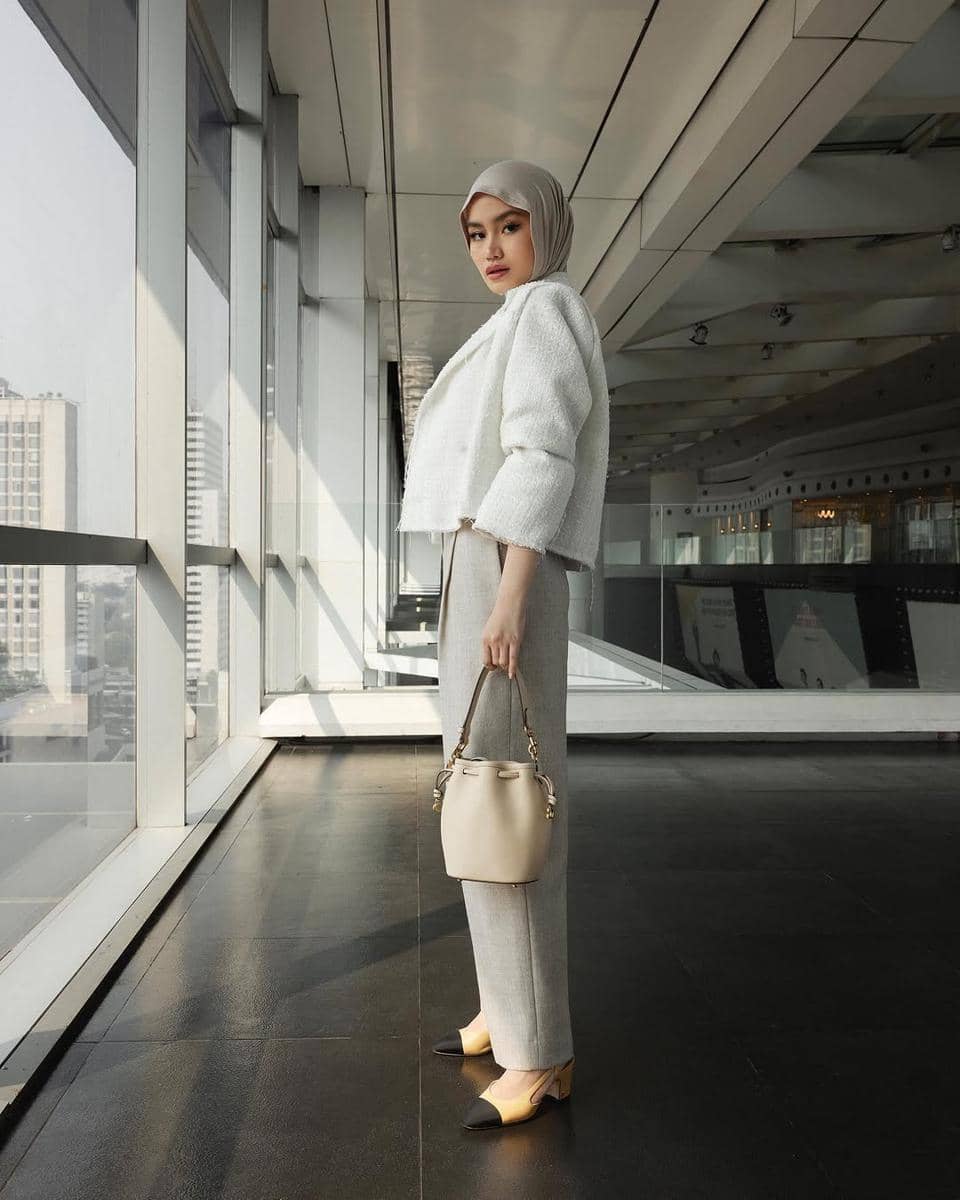 Seorang wanita berhijab mengenakan atasan putih dan celana abu-abu, memegang tas krem di area gedung modern dengan jendela besar.