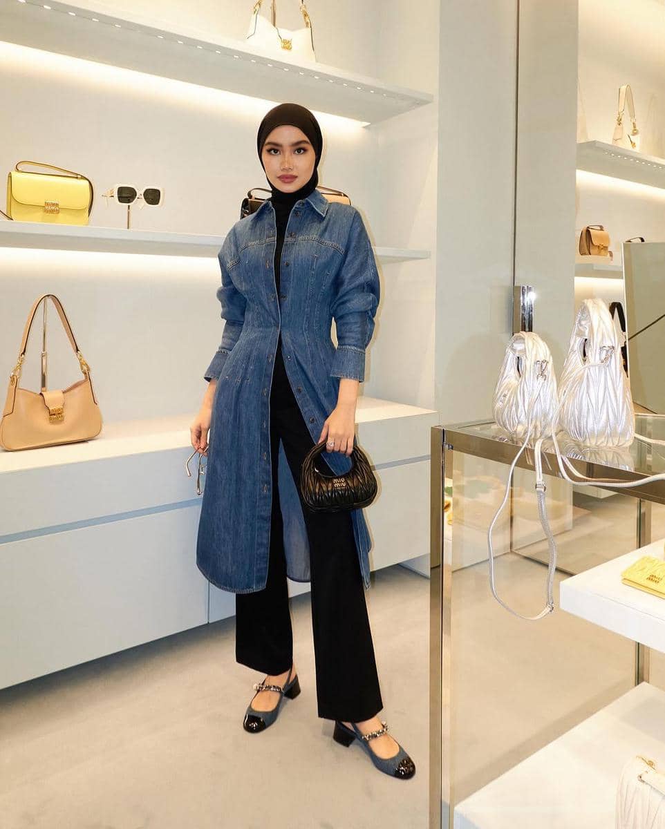 Seorang wanita berhijab mengenakan kemeja denim panjang dipadukan dengan celana hitam dan tas kecil hitam di butik fashion modern.