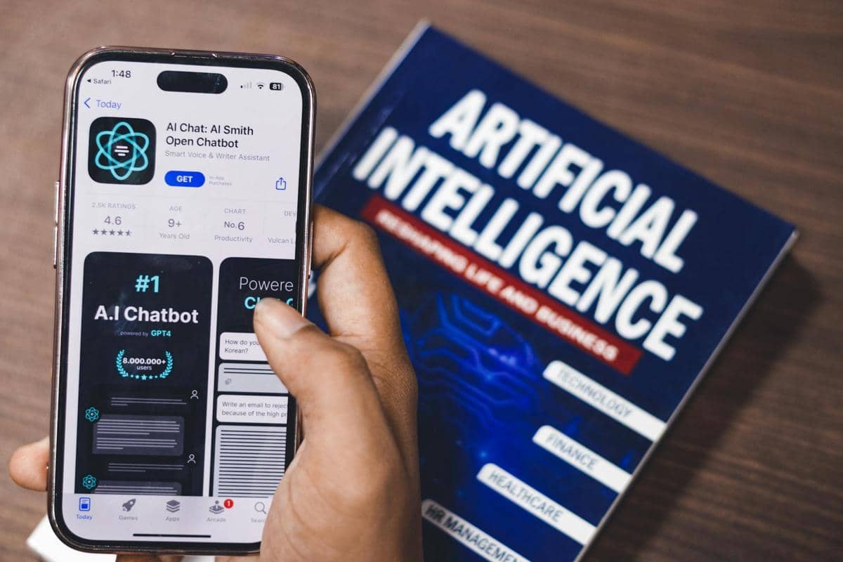 Seseorang memegang ponsel yang menampilkan aplikasi chatbot AI di samping buku bertuliskan Artificial Intelligence di atas meja kayu.