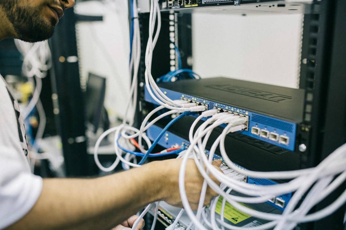 Seorang teknisi sedang mengatur kabel jaringan pada rak server di pusat data dengan perangkat jaringan berwarna biru.