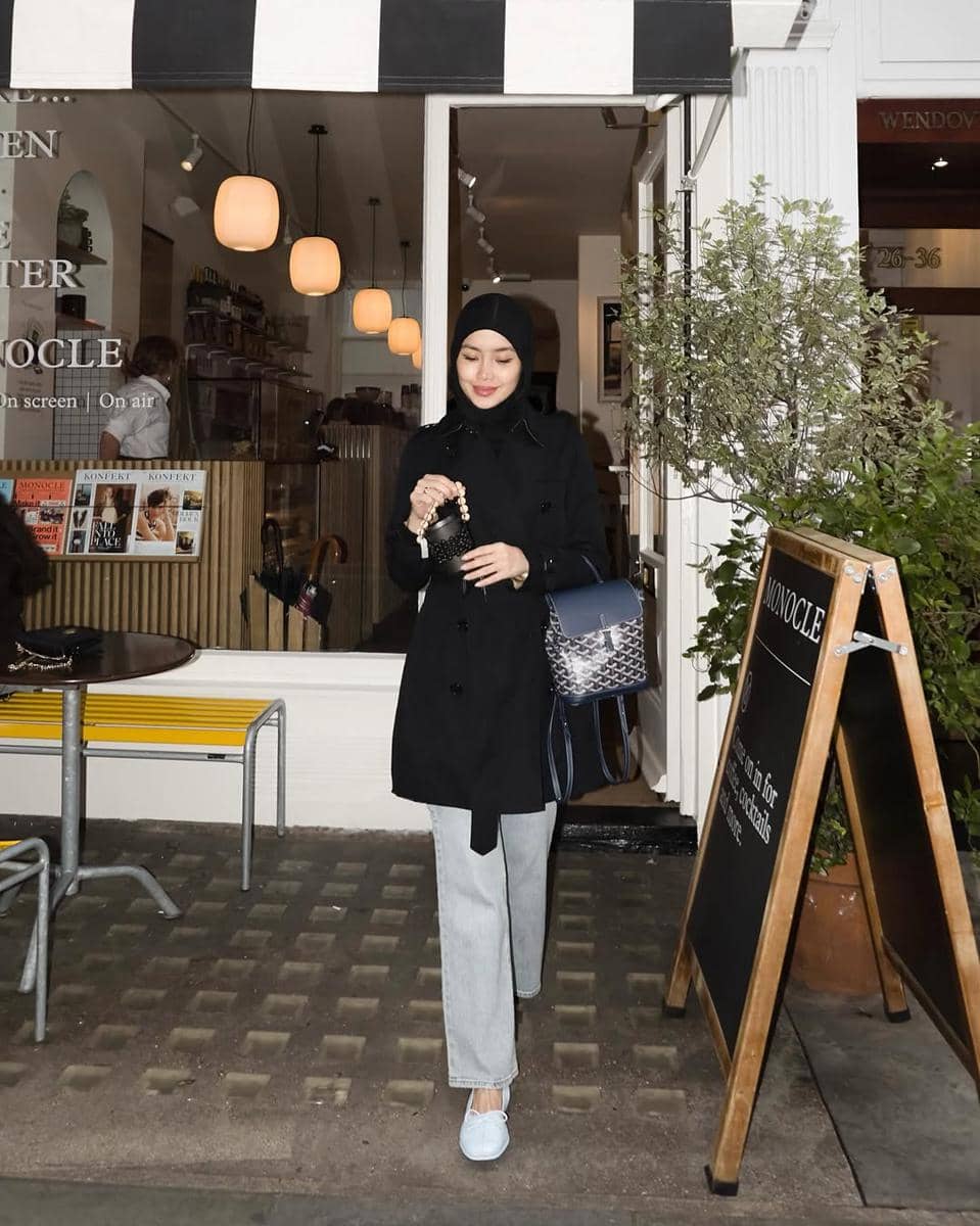 Seorang wanita berhijab mengenakan mantel hitam dan celana denim abu-abu muda sambil berdiri di depan kafe dengan dekorasi modern.