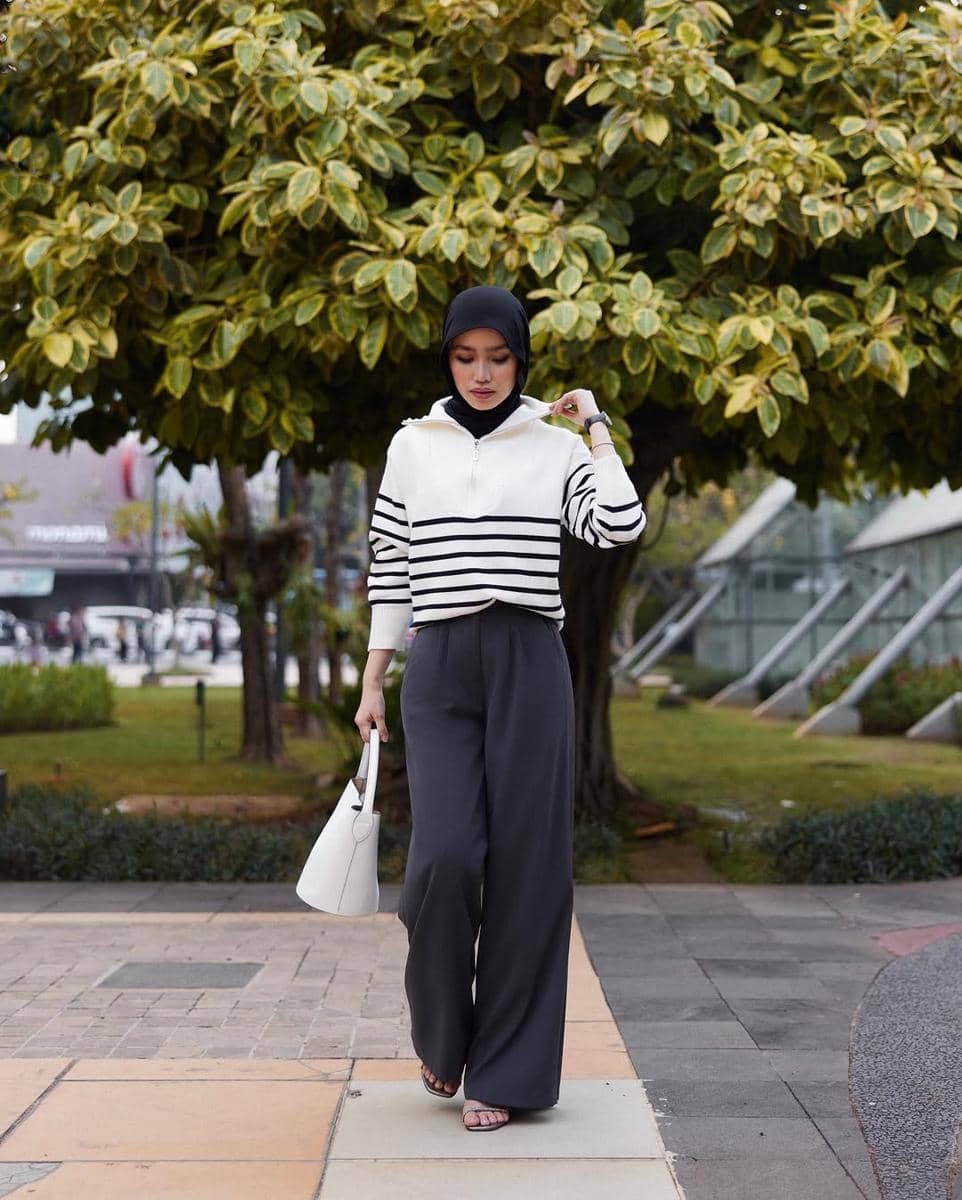 Seorang wanita berhijab mengenakan atasan putih bergaris hitam dan celana panjang lebar berwarna gelap berjalan di area taman kota.