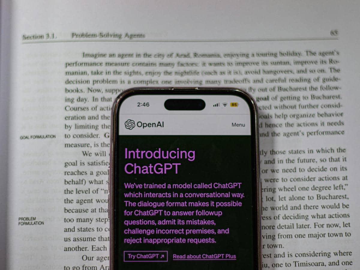 Layar ponsel menampilkan halaman OpenAI dengan teks pengenalan ChatGPT di atas buku terbuka yang berisi tulisan berbahasa Inggris.