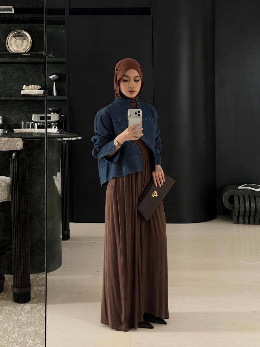 Seorang wanita berhijab cokelat mengenakan jaket denim biru dan rok panjang cokelat sambil berfoto di depan cermin dalam ruangan modern.