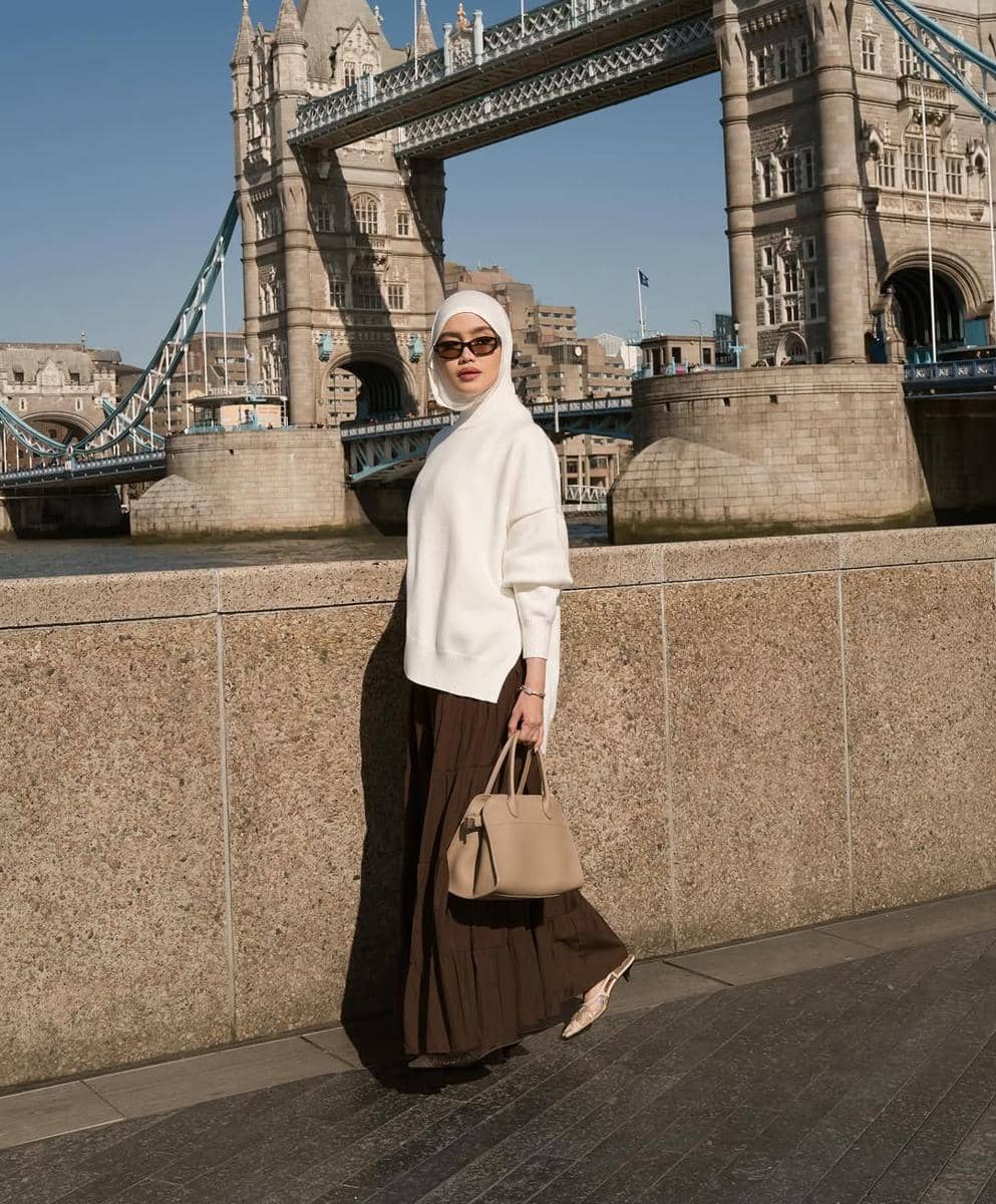Seorang wanita berhijab mengenakan atasan putih dan rok cokelat panjang sambil membawa tas krem, berpose di dekat Tower Bridge London.