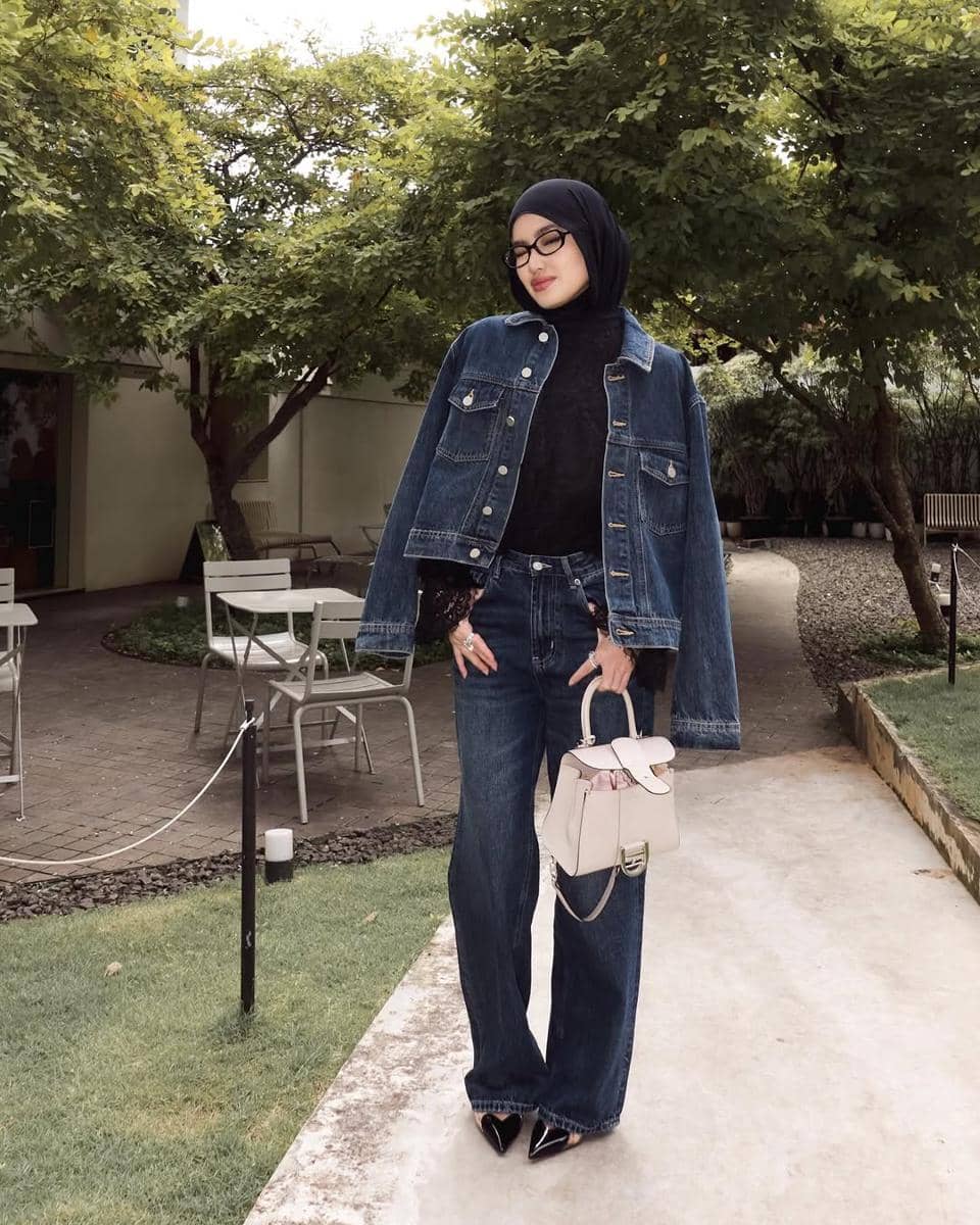Seorang wanita berhijab mengenakan jaket dan celana denim biru, memadukannya dengan atasan hitam dan tas tangan krem di area taman.