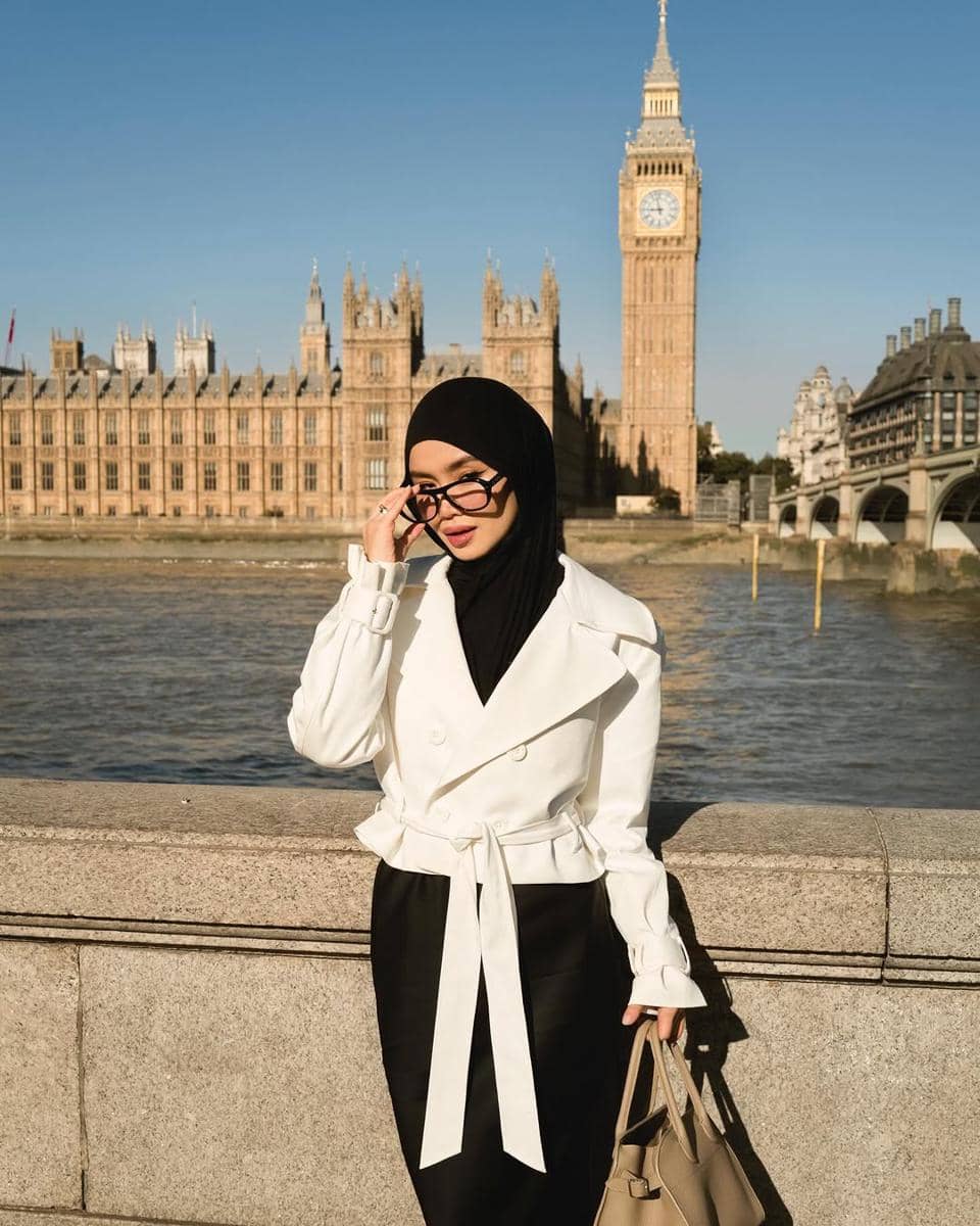 Seorang wanita berhijab hitam mengenakan atasan putih dan rok hitam berpose di depan Big Ben dan Gedung Parlemen London pada siang hari.