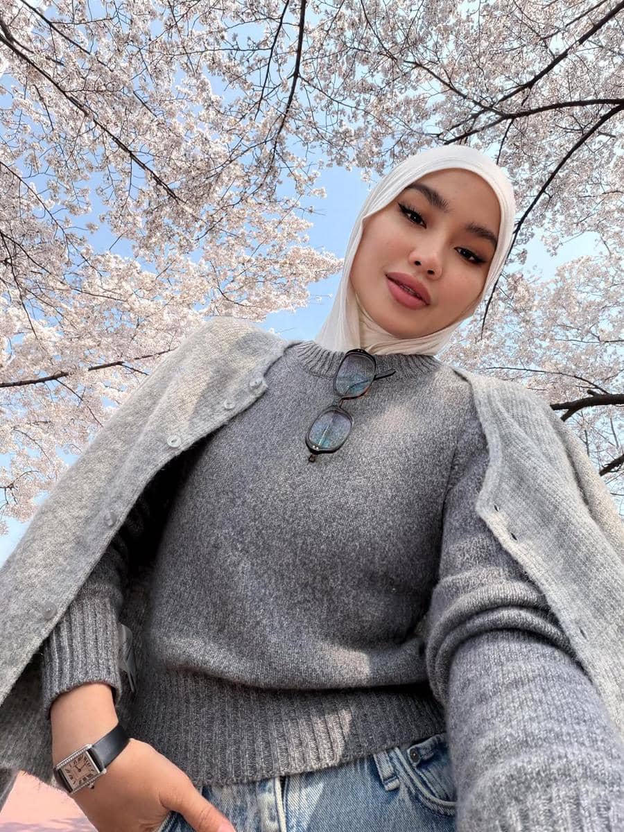 Seorang wanita berhijab mengenakan sweater abu-abu dan celana denim berpose di bawah pohon sakura bermekaran dengan langit biru cerah.
