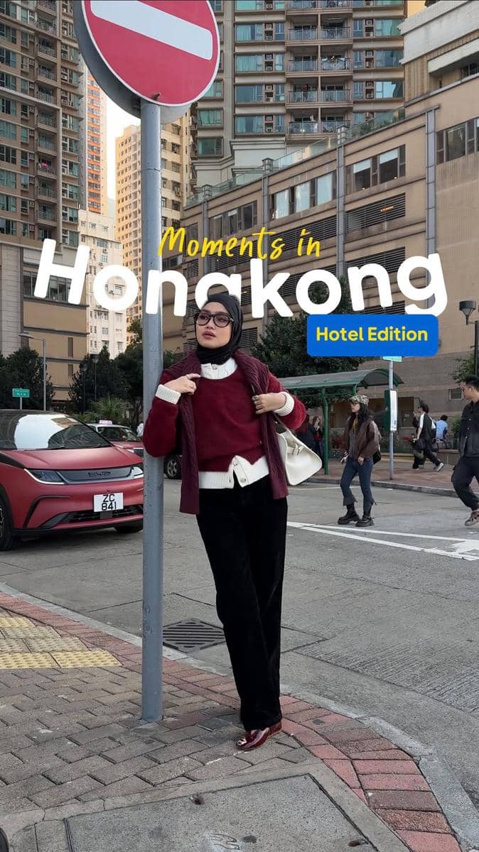 Seorang wanita berhijab bergaya dengan atasan putih dan sweater merah di jalan kota Hong Kong dengan latar gedung tinggi.