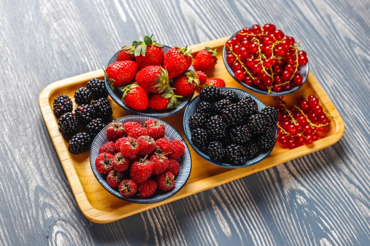 Beragam buah beri segar seperti stroberi, raspberry, blackberry, dan red currant tersaji dalam mangkuk di atas nampan kayu.