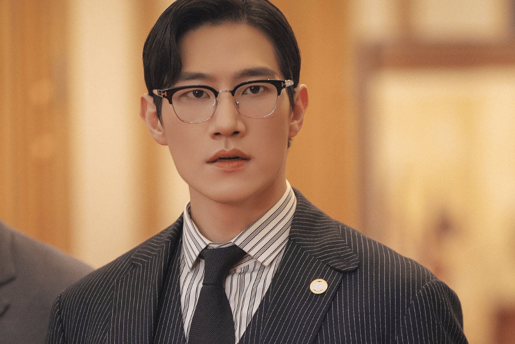 Seorang pria mengenakan setelan jas garis-garis, kemeja putih, dasi hitam, dan kacamata dalam cuplikan drama Korea Perfect Crown.