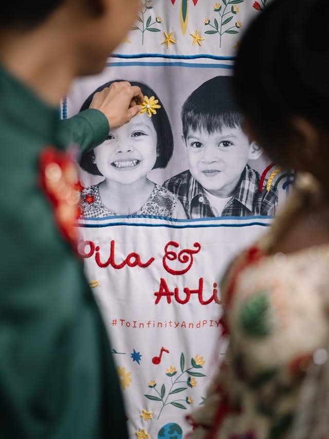 Pasangan mengenakan busana tradisional menatap kain bergambar dua anak kecil dengan tulisan nama dan hiasan bunga berwarna cerah.