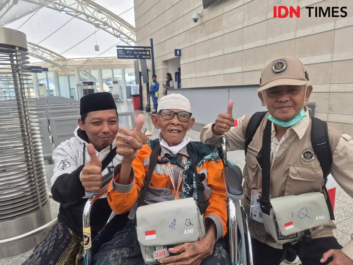 Seorang jemaah haji lanjut usia duduk di kursi roda bersama dua pendampingnya sambil tersenyum dan mengacungkan jempol di Bandara Madinah.