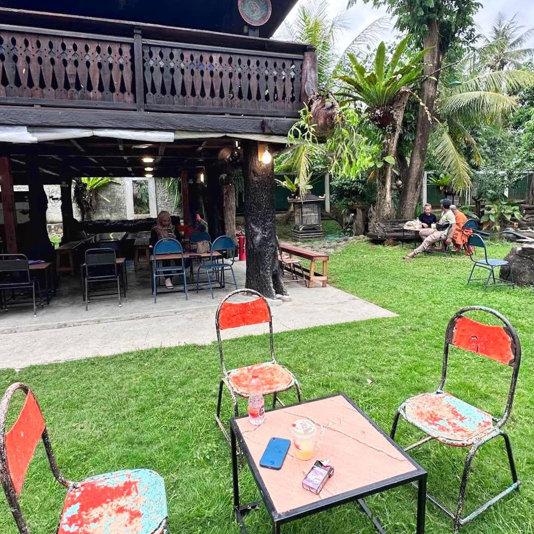 Area duduk outdoor Kopi Merbaoe di Bandar Lampung dengan kursi besi berwarna merah dan biru di halaman berumput hijau.