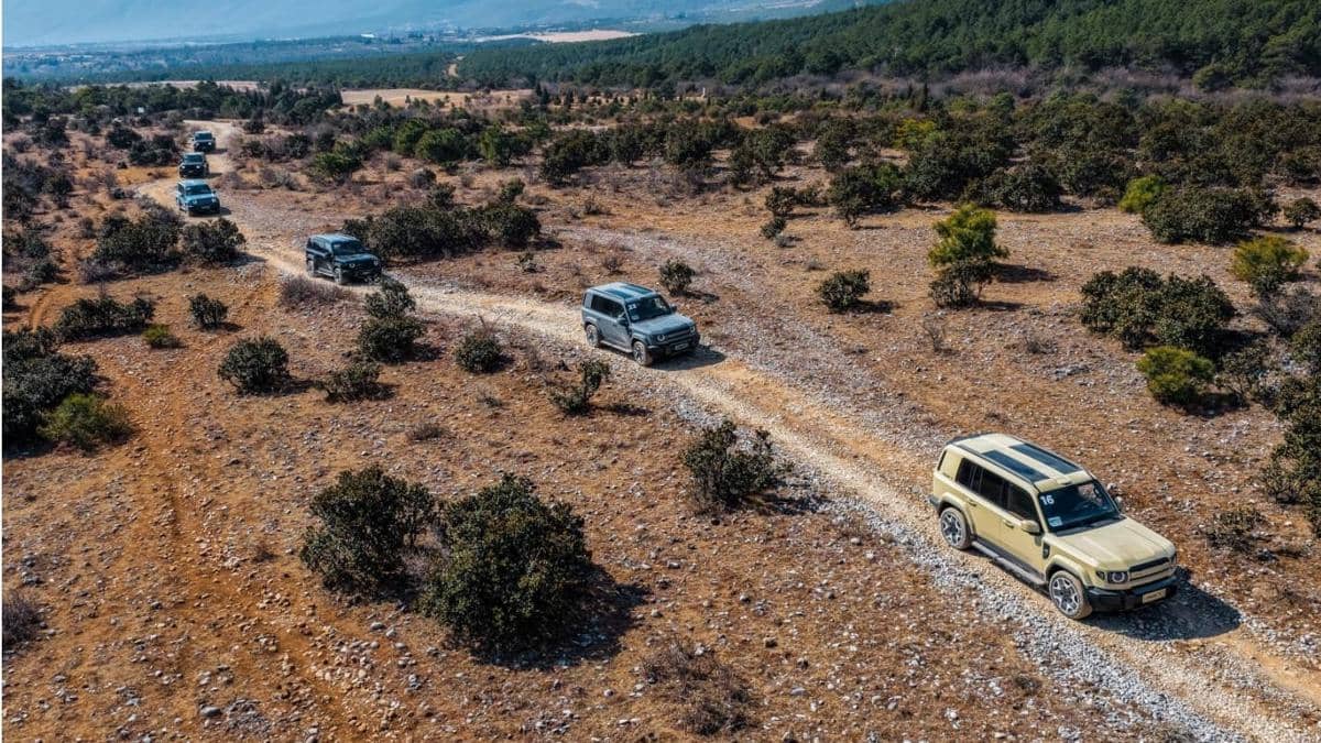 Beberapa mobil SUV melintasi jalur berbatu di area perbukitan kering Wuhu, China, dengan latar belakang hutan dan pegunungan.