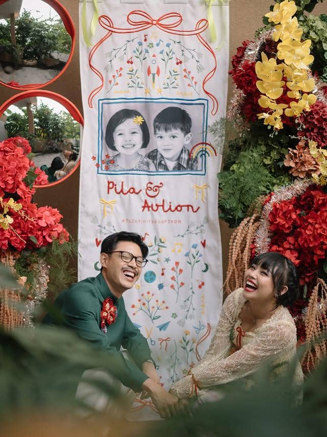 Pasangan Aulion dan Pila tertawa bahagia saat acara lamaran dengan latar dekorasi bunga warna-warni dan banner bergambar masa kecil mereka.
