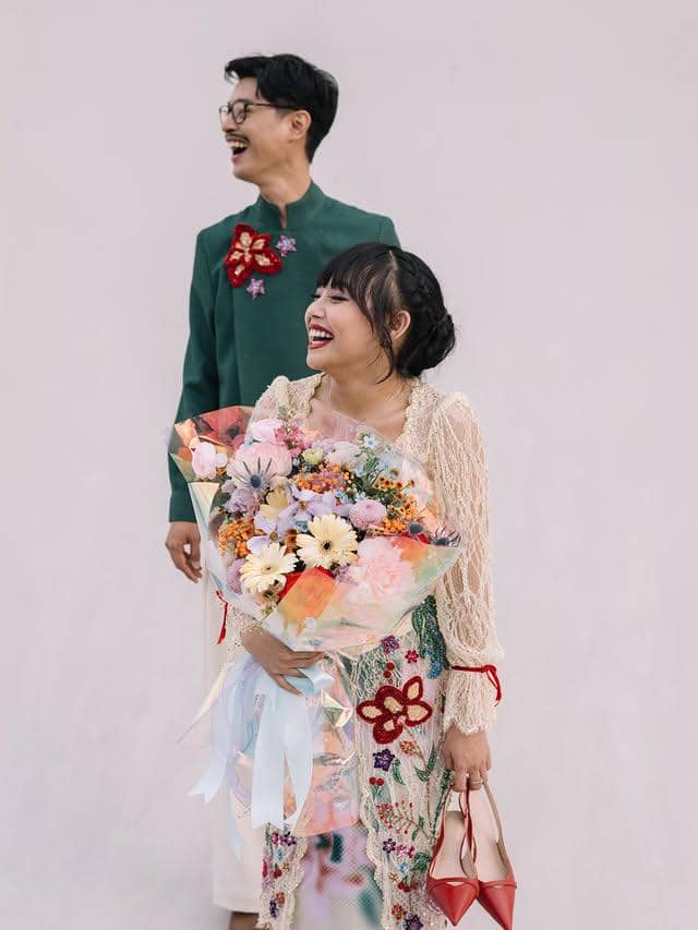Sepasang pria dan wanita tersenyum bahagia, wanita memegang buket bunga warna-warni dan tas merah, mengenakan kebaya modern.
