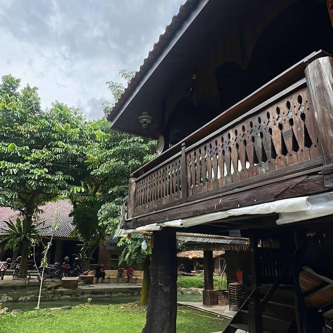 Bangunan kayu tradisional dengan balkon ukiran di Kopi Merbaoe Bandar Lampung, dikelilingi pepohonan hijau dan area duduk luar ruangan.