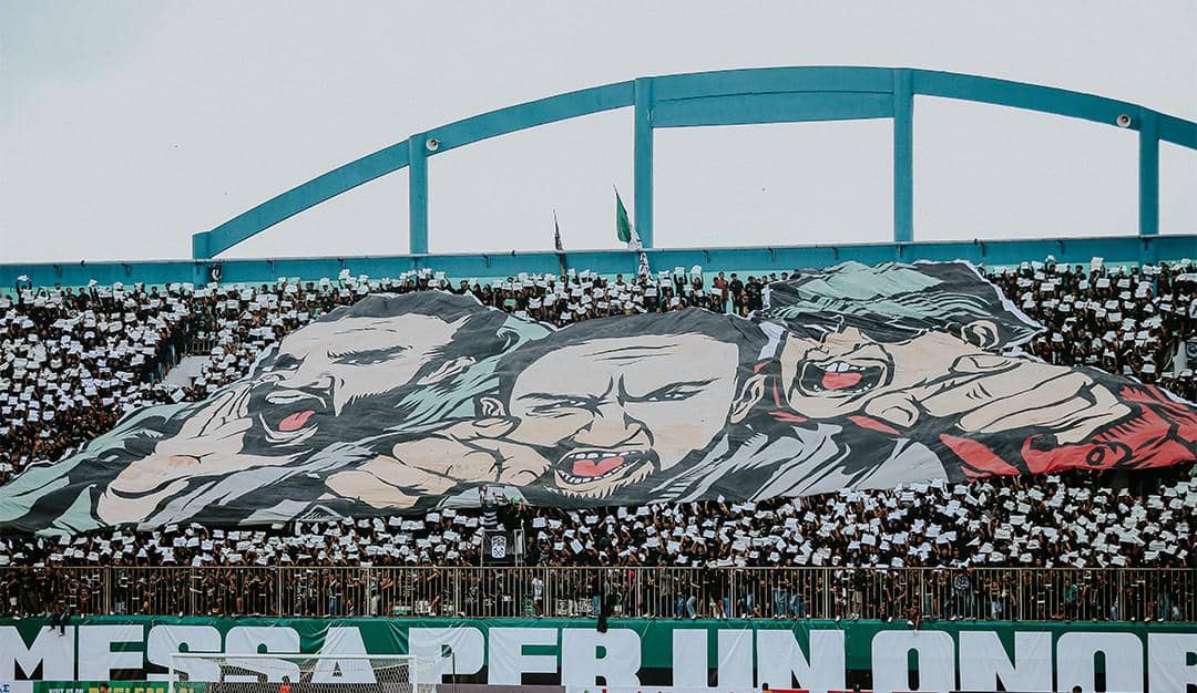 Suporter PSS Sleman Brigata Curva Sud menampilkan koreografi besar bergambar tiga wajah di tribun stadion dengan latar warna hijau dan putih.